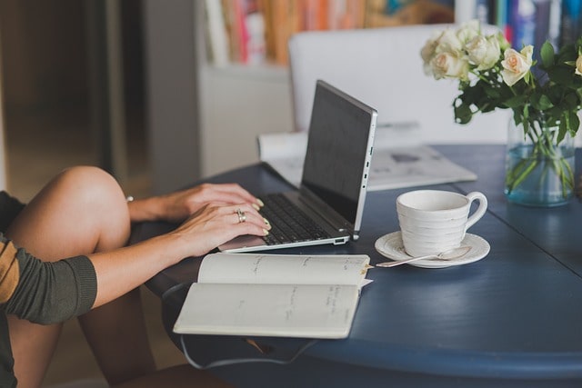 una persona estudia en su laptop. hay una libreta abierta al lado del laptop y una taza de café y un jarrón con flores en la mesa