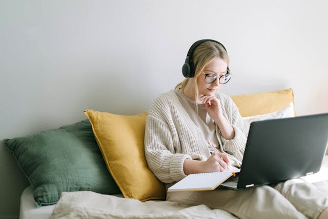 una mujer sentada en su cama con unos auriculares puestos y un laptop y un cuaderno en sus piernas. parece que está haciendo un listening en inglés mientras toma notas de lo que escucha